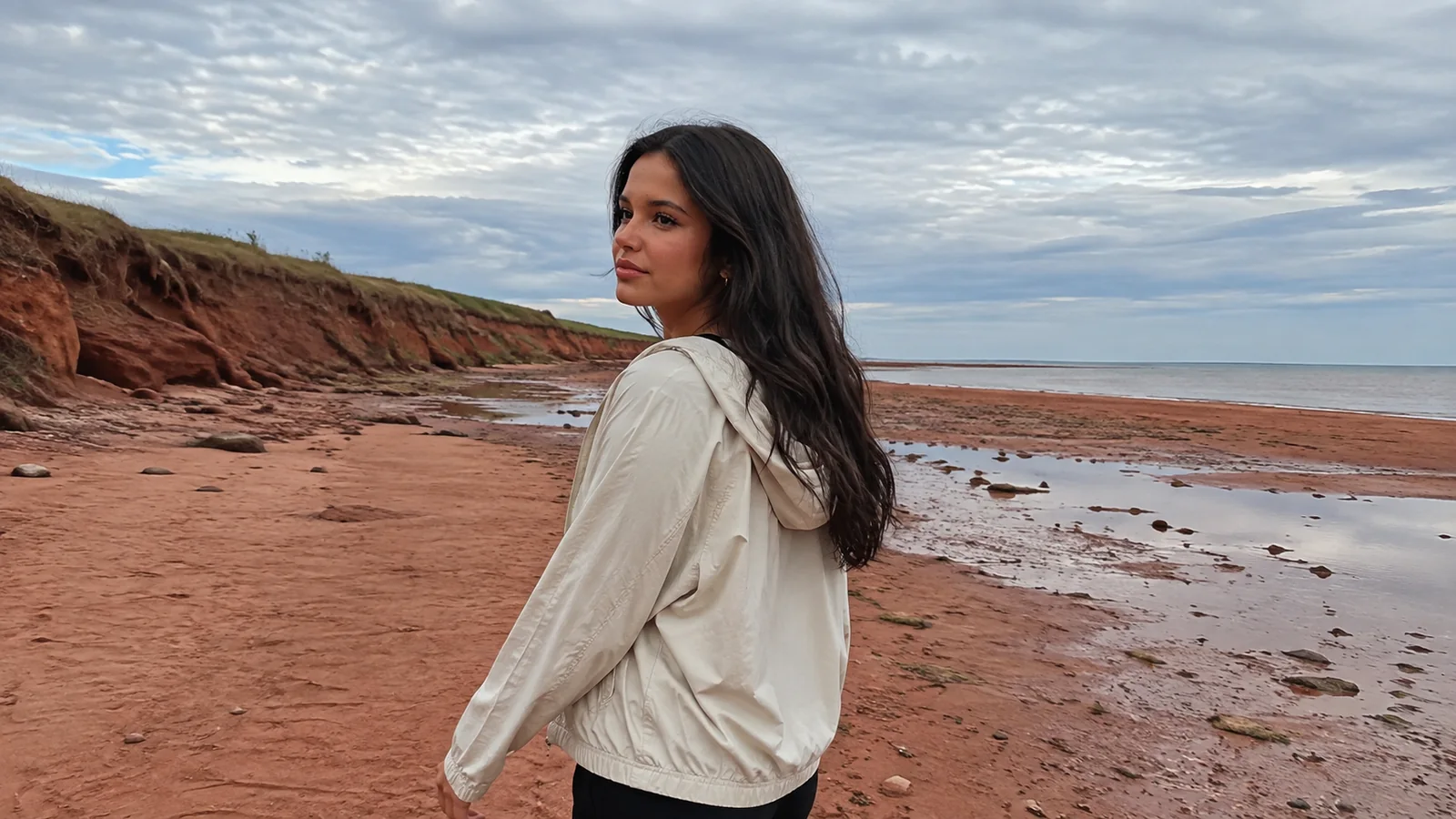 Woman walking beside red sandstone cliffs and shallow shoreline pools.