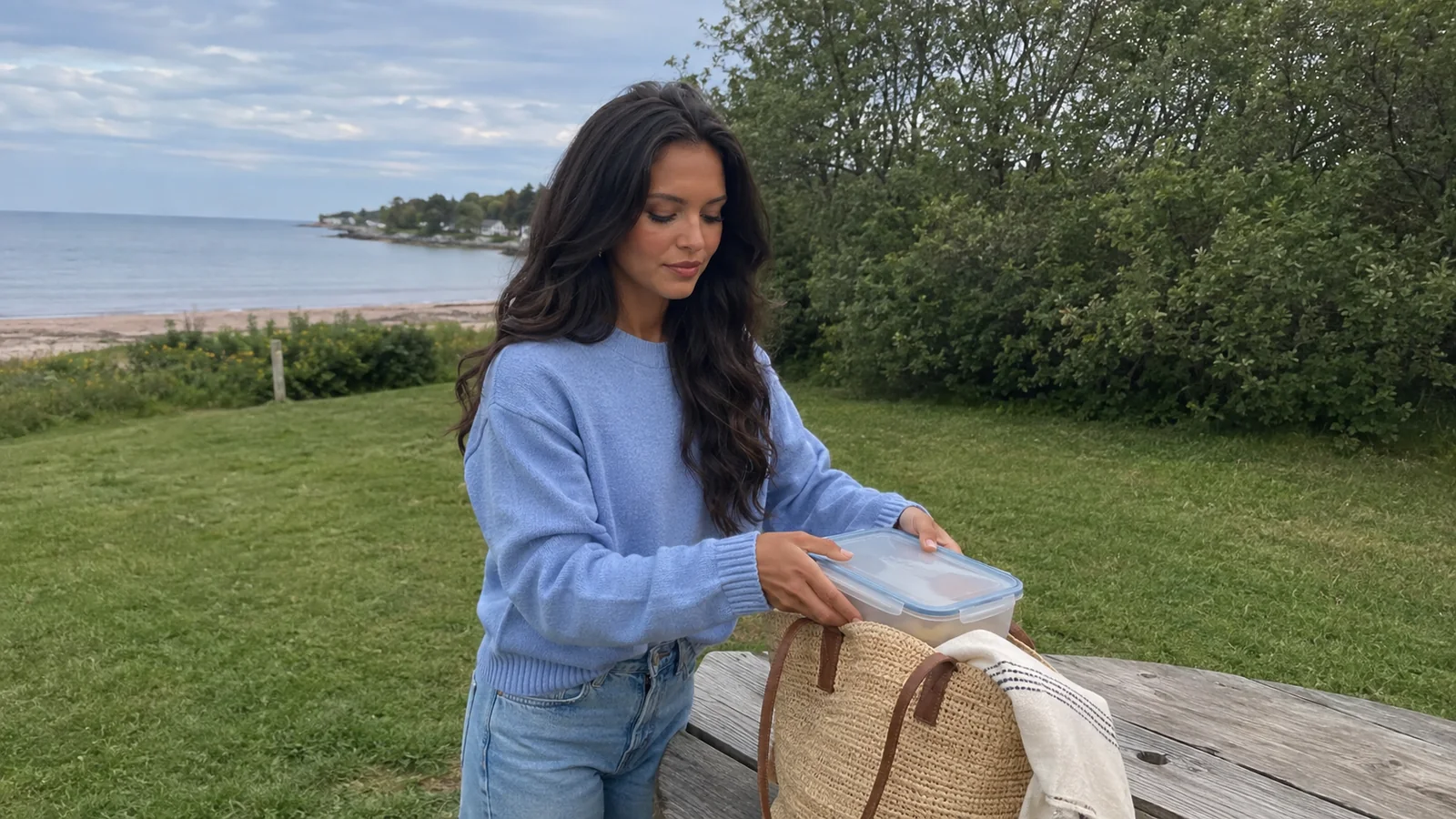 Woman placing a food container into a tote at a waterfront picnic table.