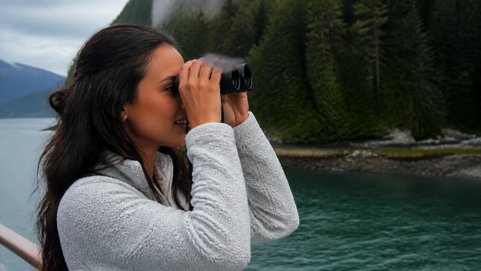 Woman using binoculars beside a forested fjord