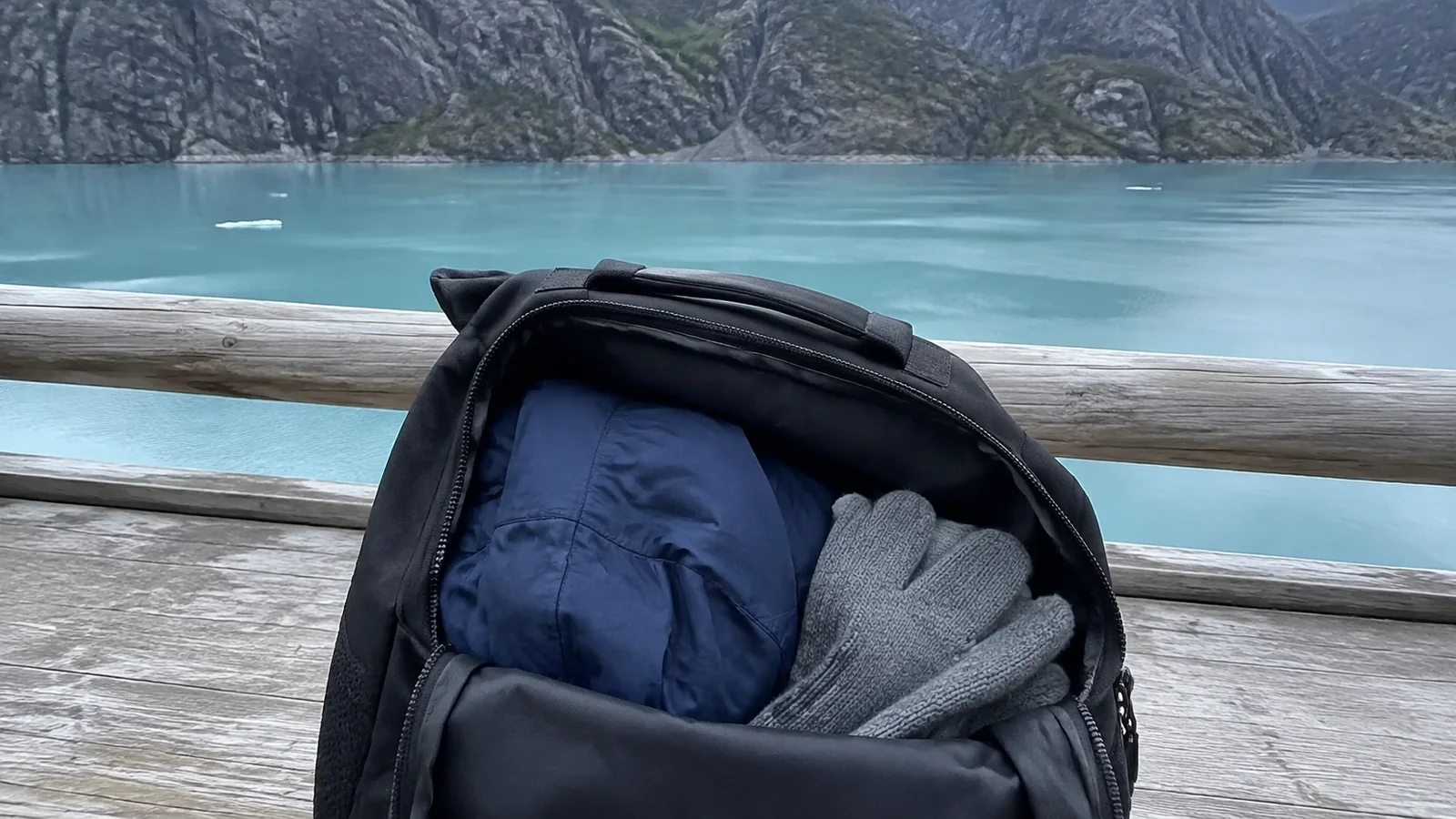 Backpack containing a jacket and gloves beside turquoise glacial water