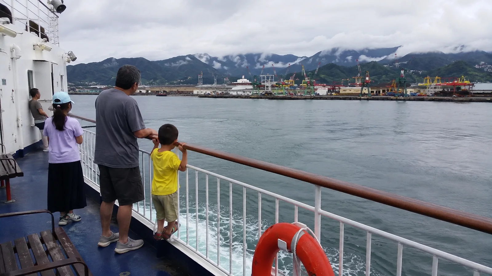 Family watching a port and mountain shoreline from a ship deck.