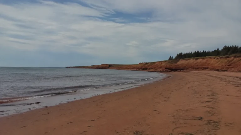 Quiet sandy beach curves toward low red cliffs beneath clouds.