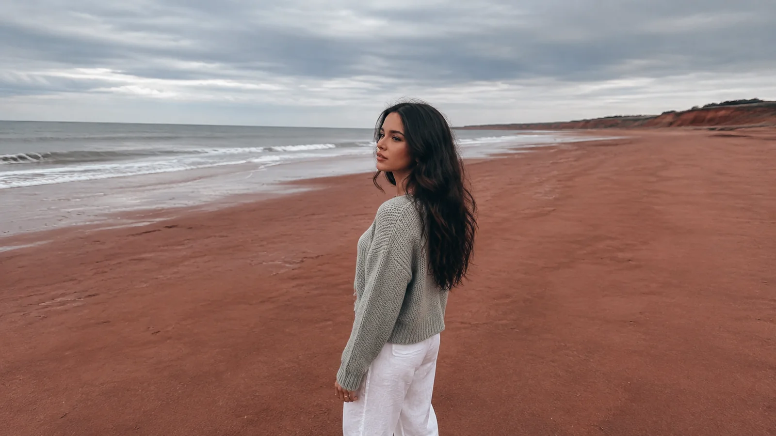 Woman standing on a red sand beach beside breaking waves.