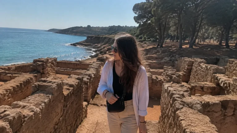Woman explores low stone ruins beside a blue sea.