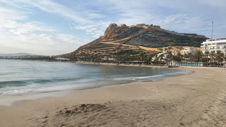 Sandy beach beside calm water beneath a rocky hill topped with stone fortifications.