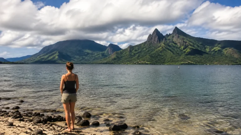 Woman standing at a rocky shoreline facing a green mountainous island.