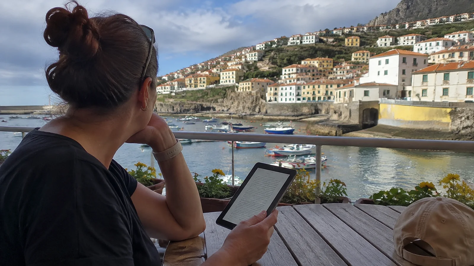 Woman reading an e-reader at an outdoor table overlooking a harbor
