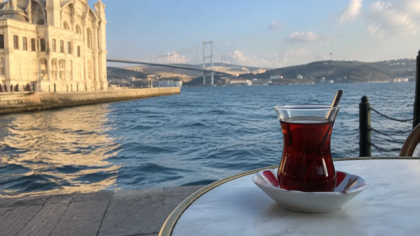 Glass of tea on a waterfront table overlooking a bridge and shoreline buildings.