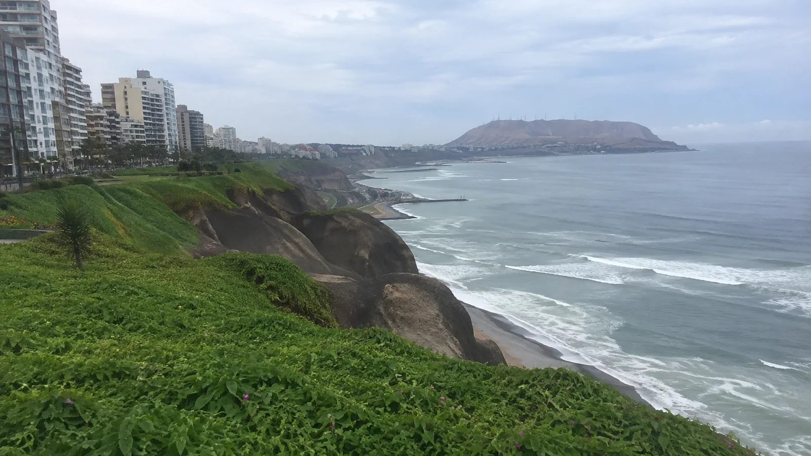Green coastal cliffs and city buildings overlook breaking waves.