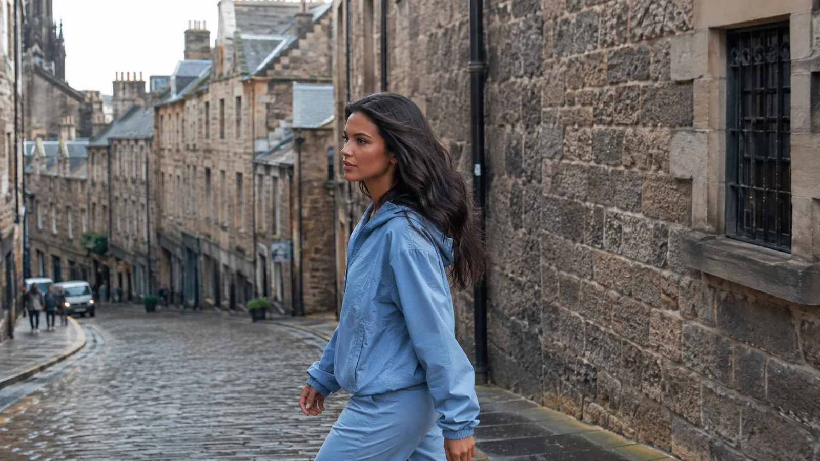 Woman stands on a cobbled street beside old stone buildings.