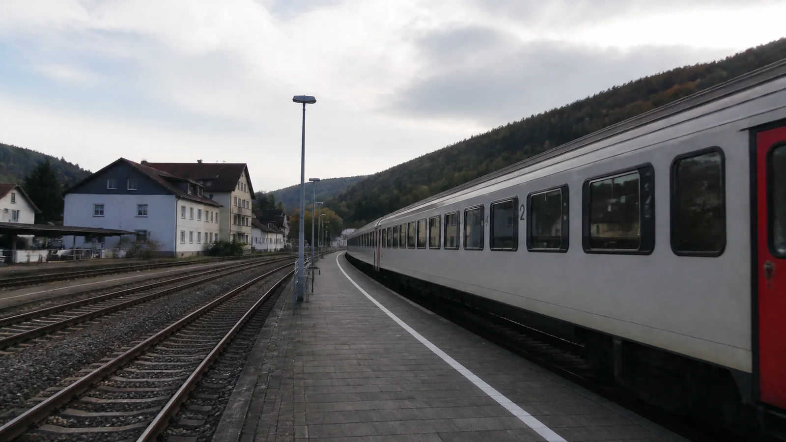 Passenger train stands beside a platform near small hillside buildings.