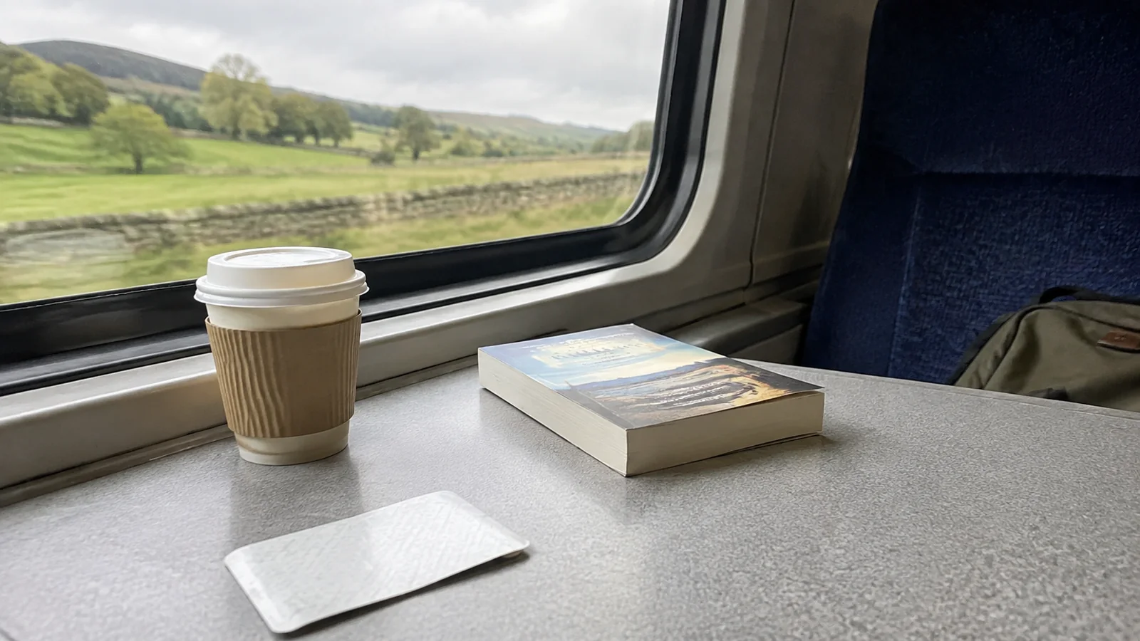 Takeaway coffee and a paperback on a train table beside green countryside.