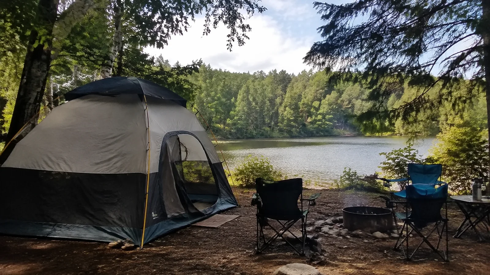 Tent and camping chairs beside a wooded lake.