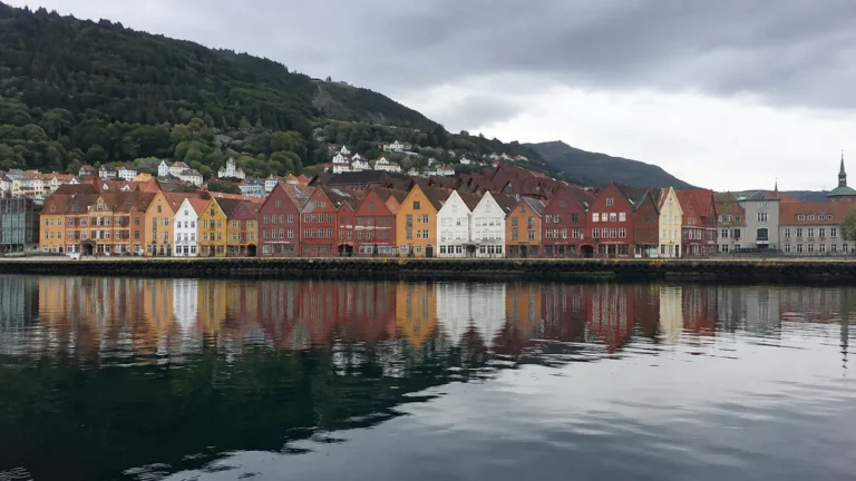 Colorful waterfront buildings reflect in calm water below a wooded hillside.