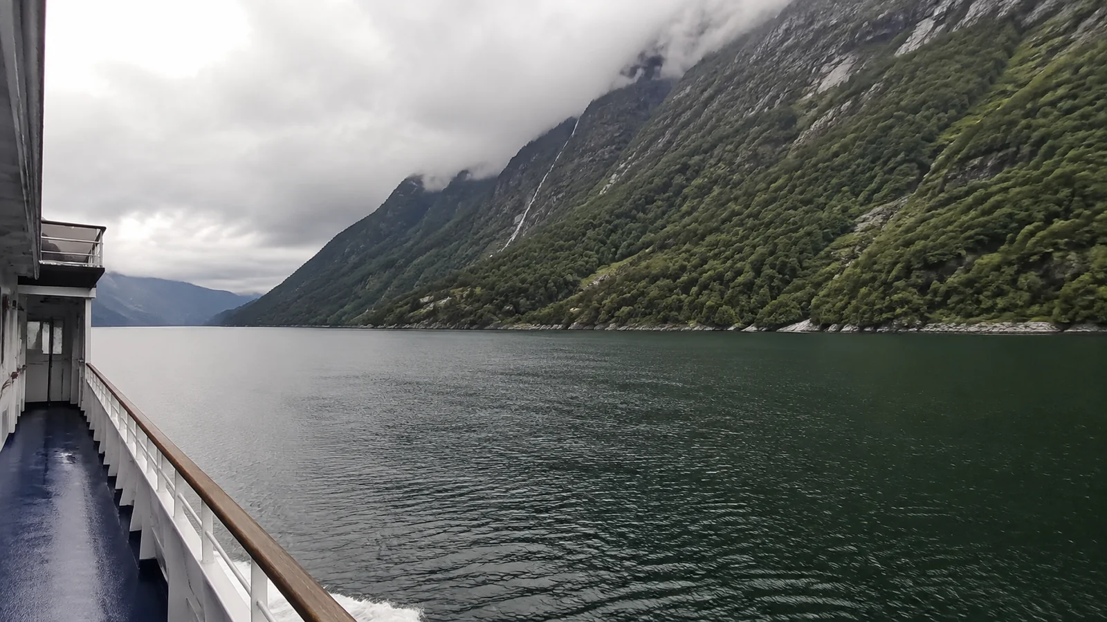 Ship deck beside a narrow fjord beneath towering green mountains.