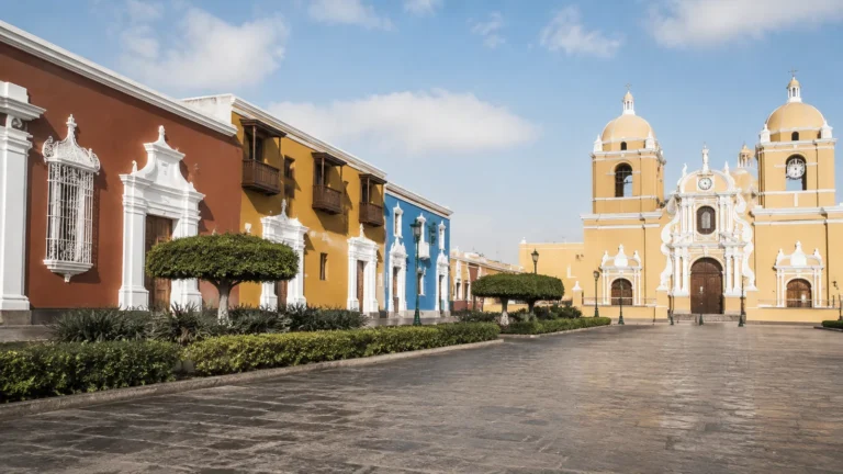 Colorful historic buildings and a yellow twin-towered church bordering a paved square.