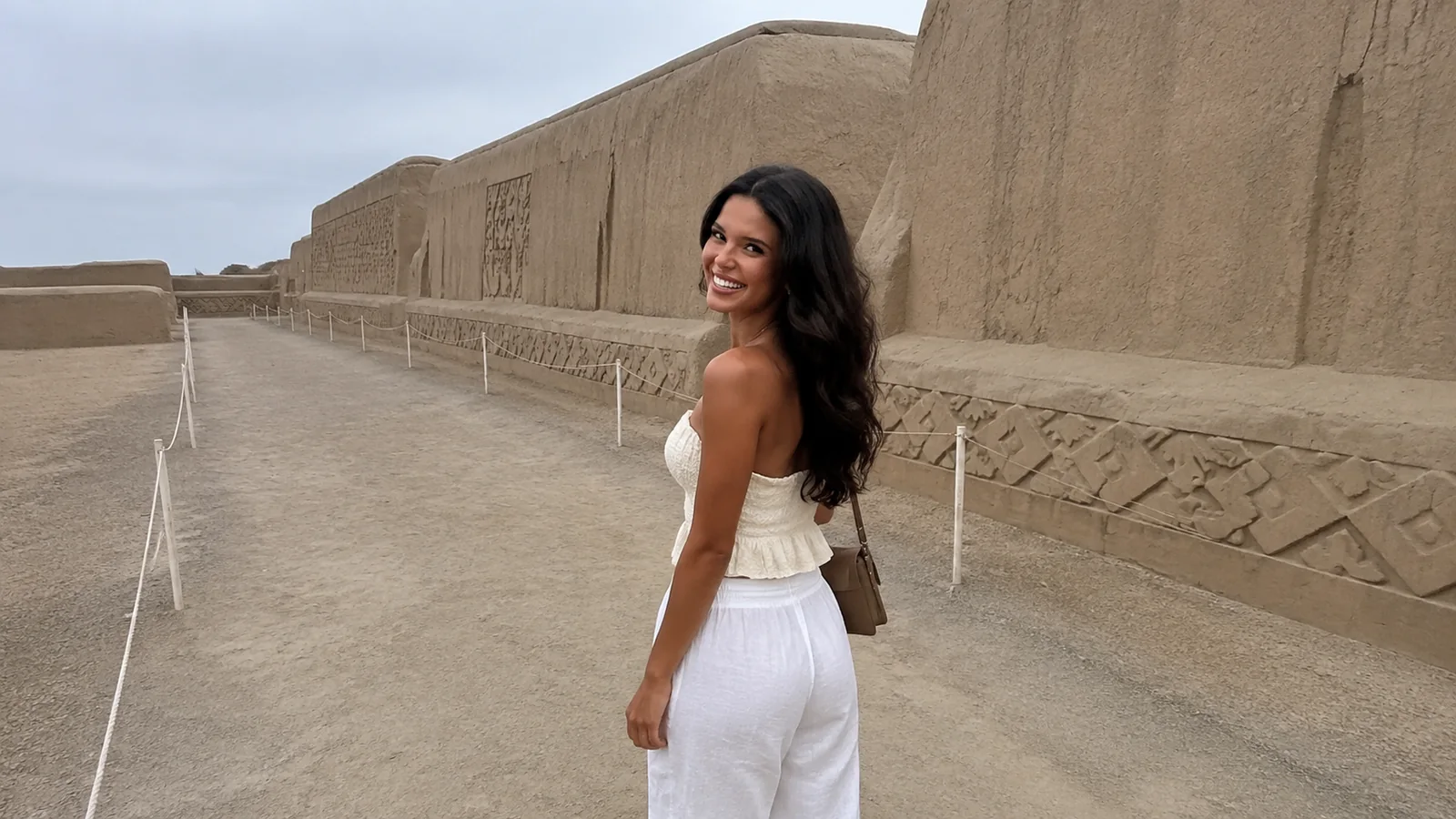 Woman in a white dress walking beside carved earthen archaeological walls.