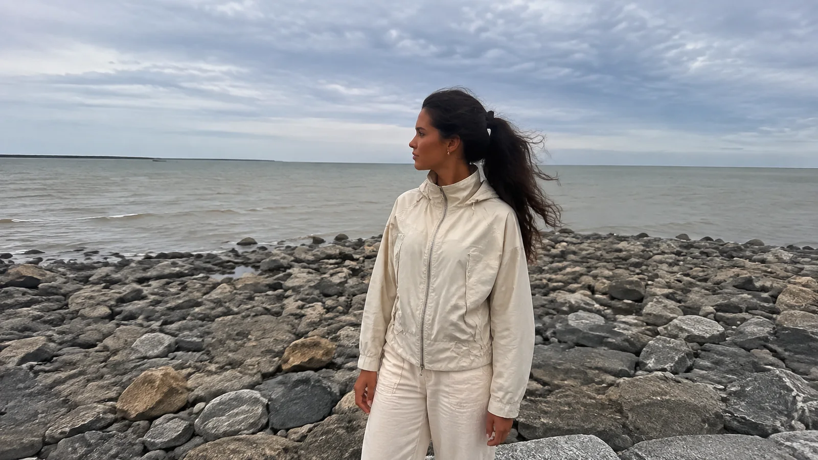 Woman in a light rain jacket stands on a rocky shore.