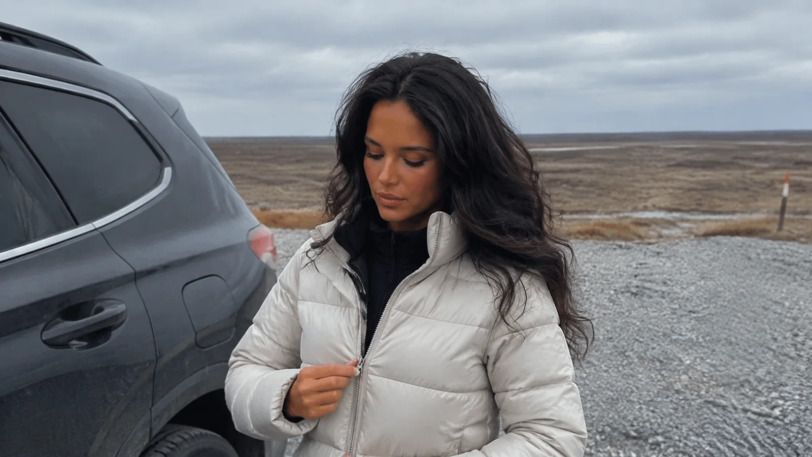 Woman adjusts her insulated jacket beside a vehicle on a gravel road.