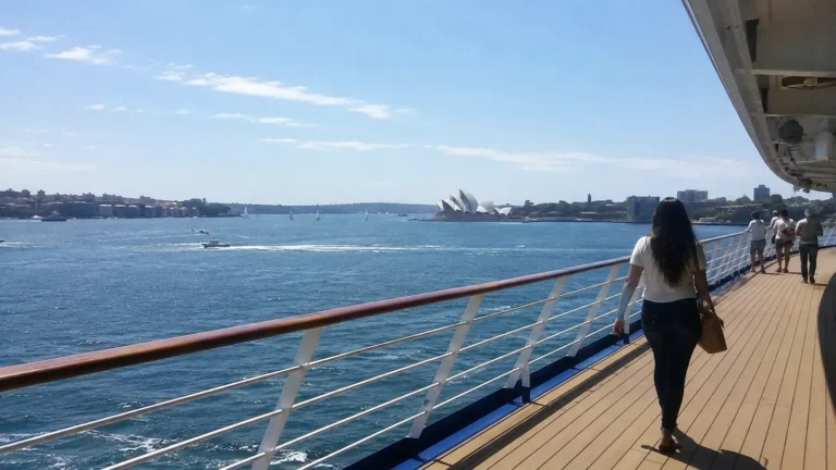Passengers walking along a ship's outer deck overlooking a busy harbor.