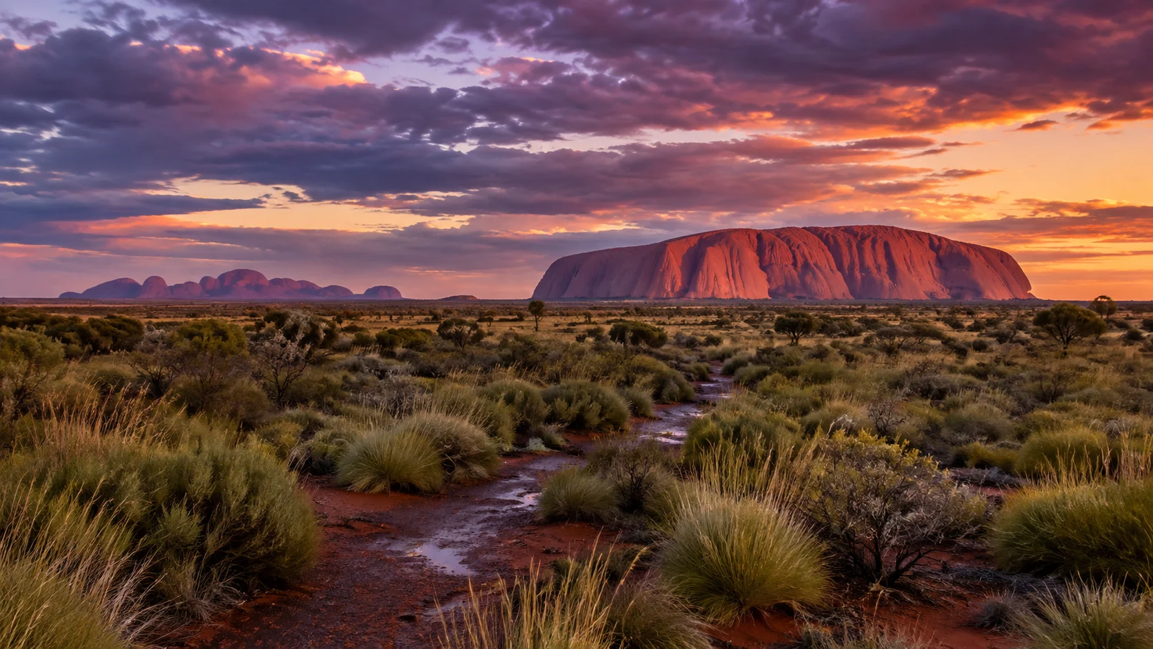 Uluṟu and distant Kata Tjuṯa glow after rain beneath a dramatic desert sunset.