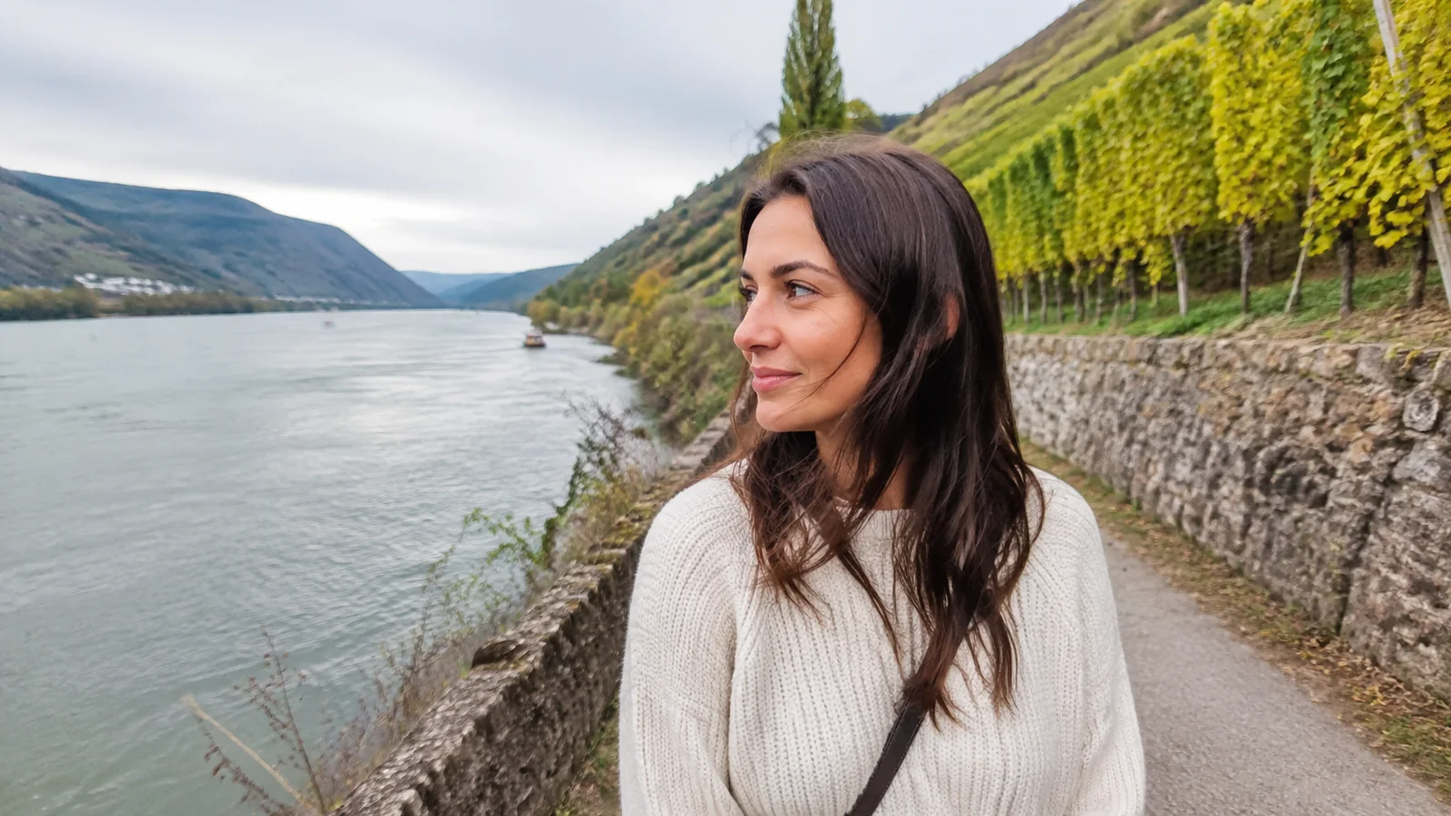 Woman walking beside a river below terraced vineyards.