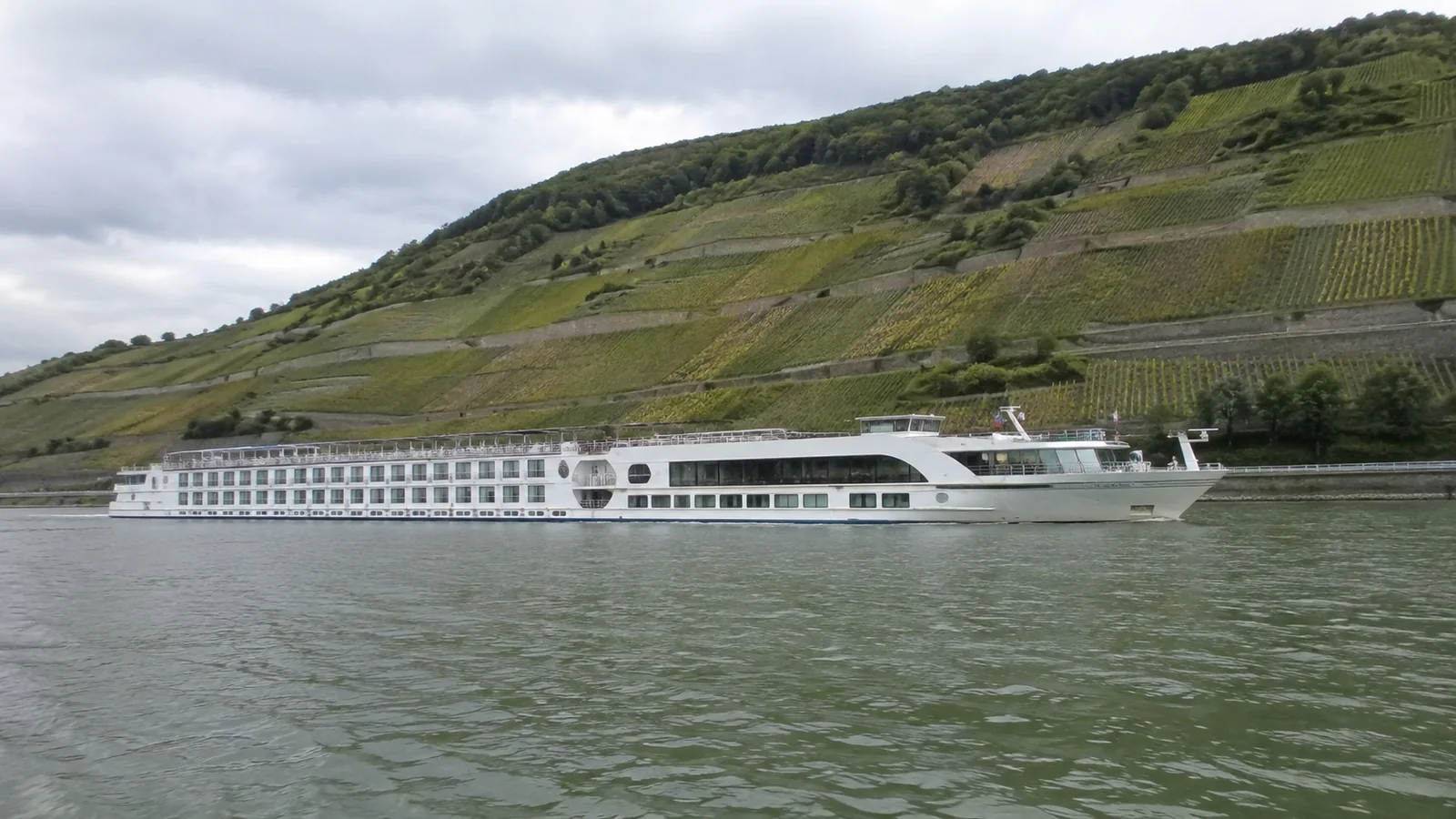 Long river cruise vessel passing below a vineyard-covered hillside.