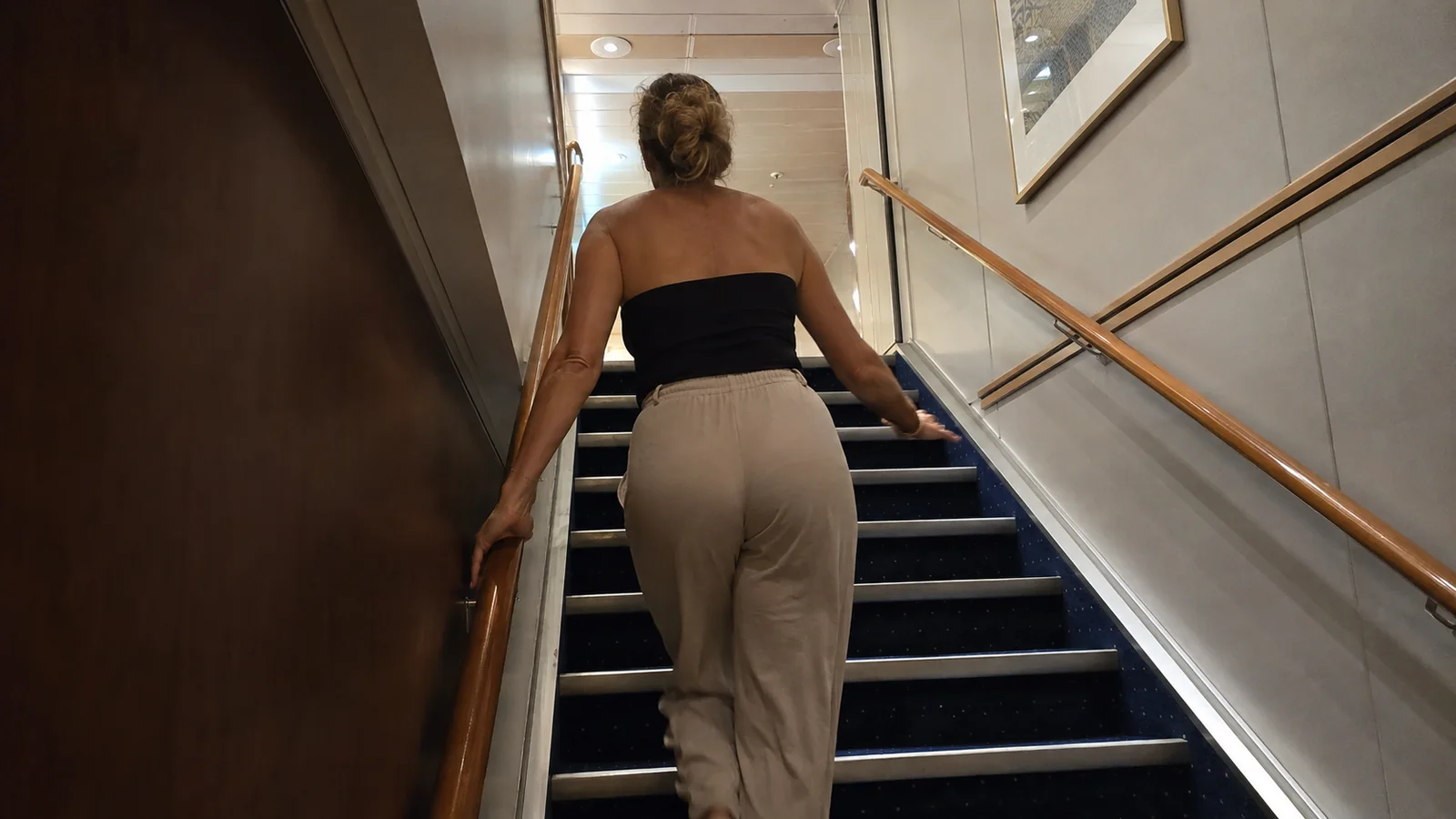 Woman climbs an interior ship staircase beside a wooden handrail.