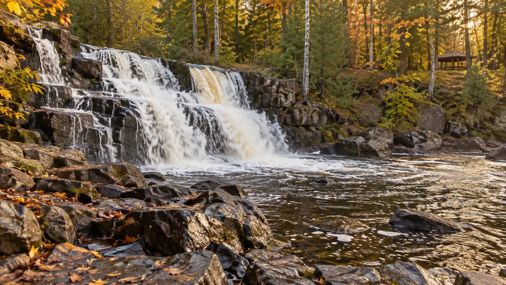 Upson Falls cascading over dark ledges beside autumn trees and a distant park shelter in Iron County.