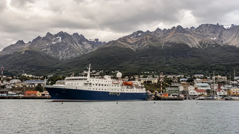 Passenger ship docked before a town backed by jagged mountains.