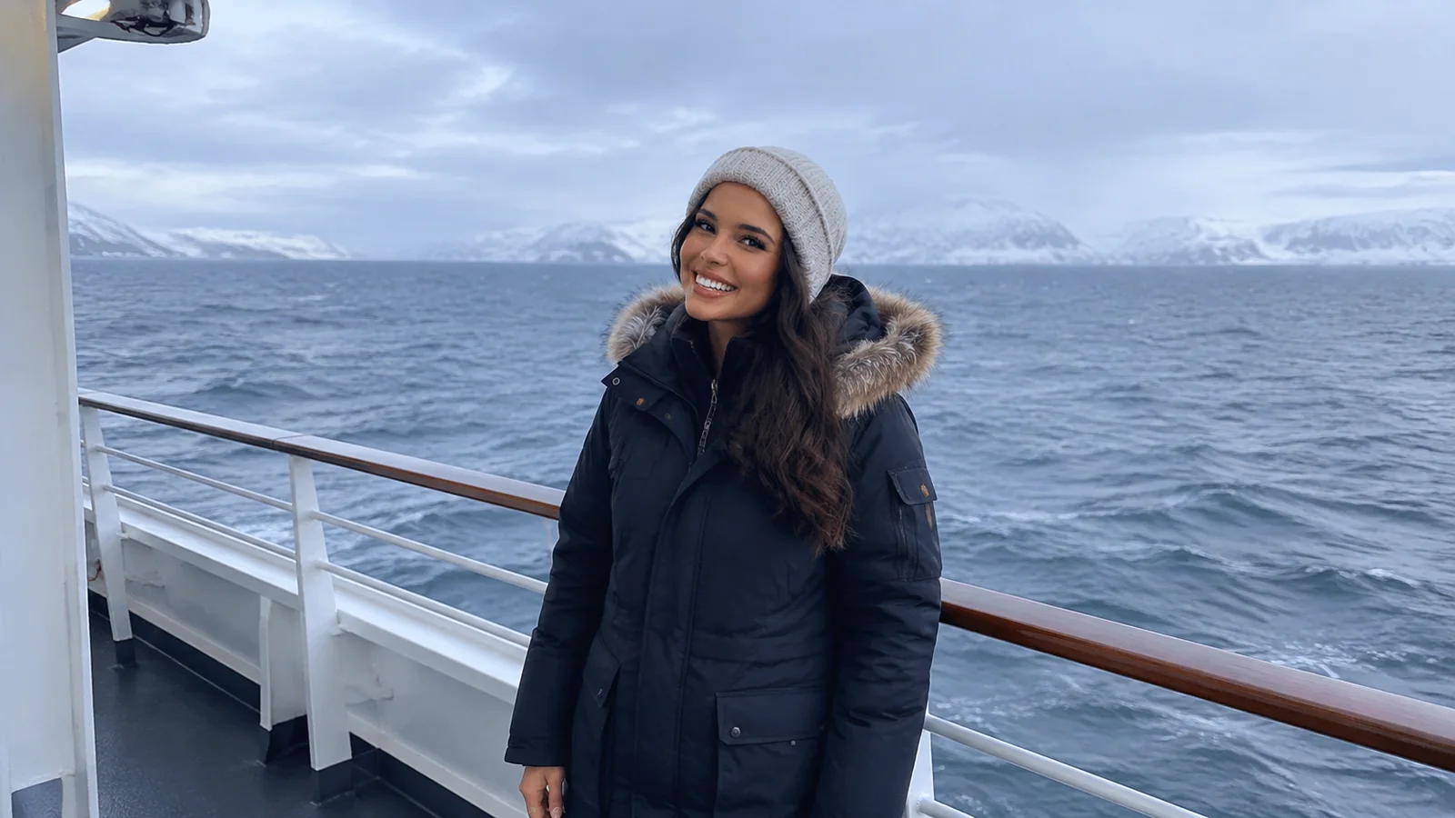 Woman wearing a winter coat on a ship deck near snowy mountains.