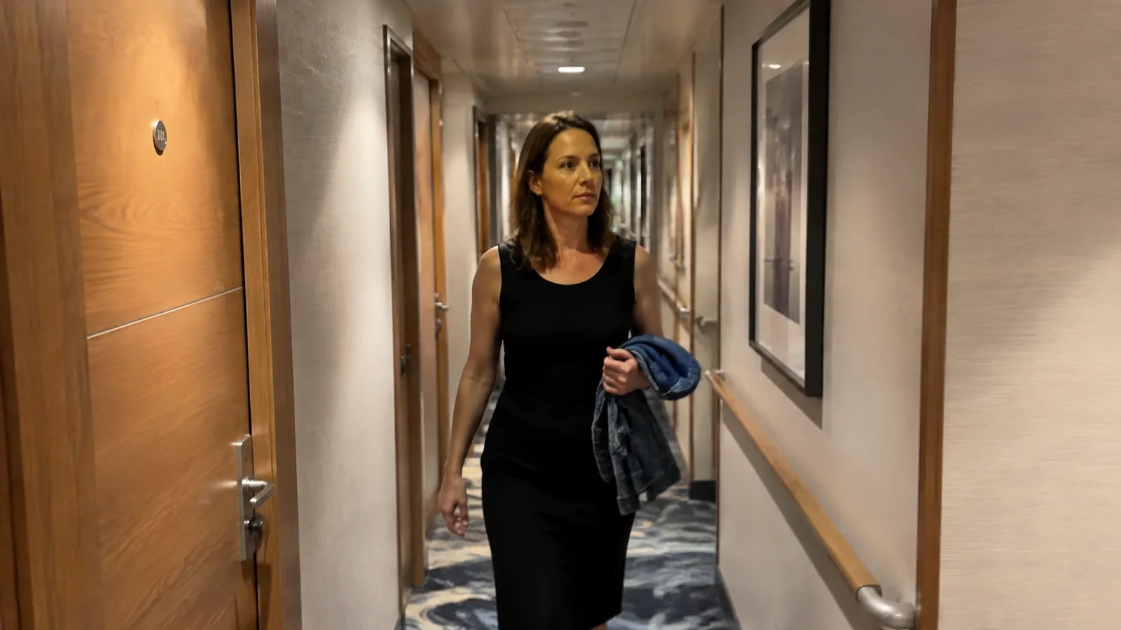 Woman carries a jacket along a carpeted ship corridor.
