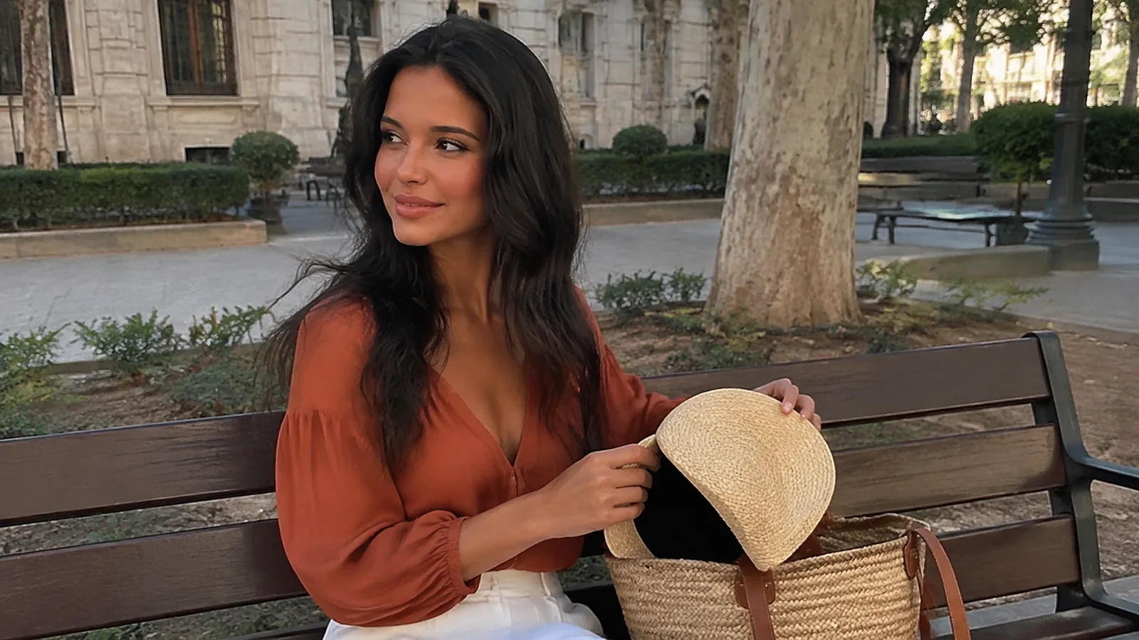 Woman holding a sunhat on a bench in a shaded square.