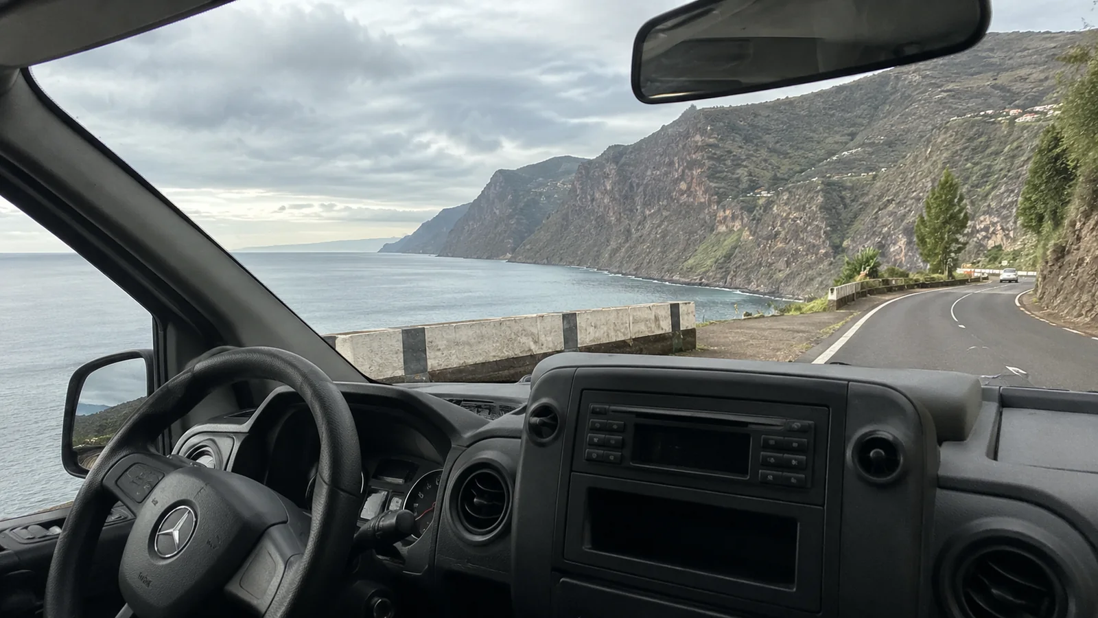 View through a van windshield along a winding coastal road beside steep cliffs and the ocean