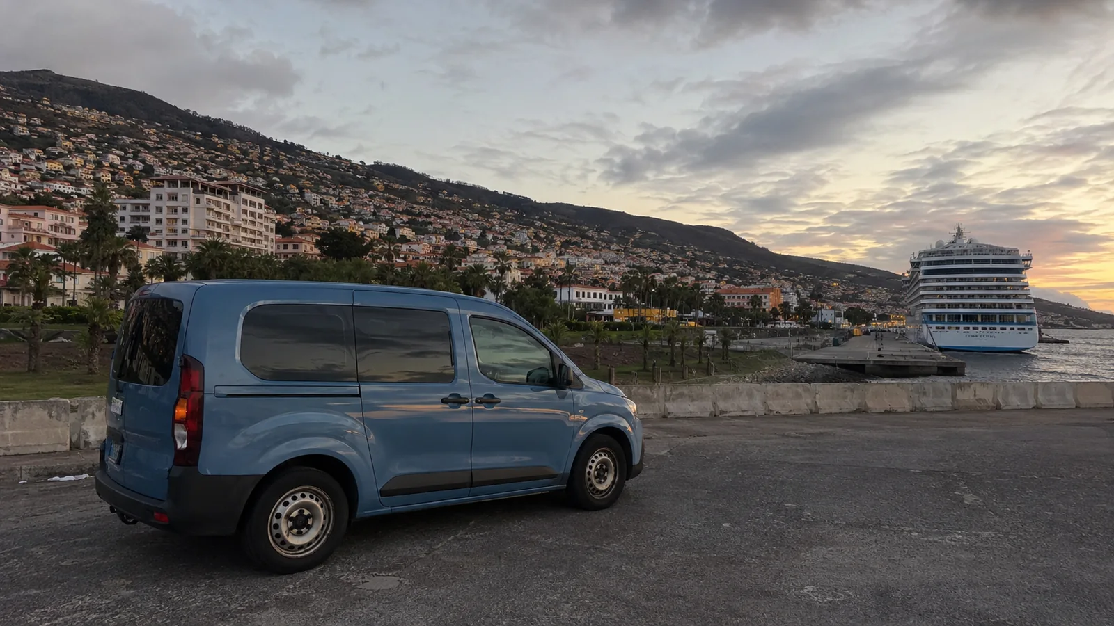 Blue passenger van parked at a waterfront with a cruise ship and hillside town behind it