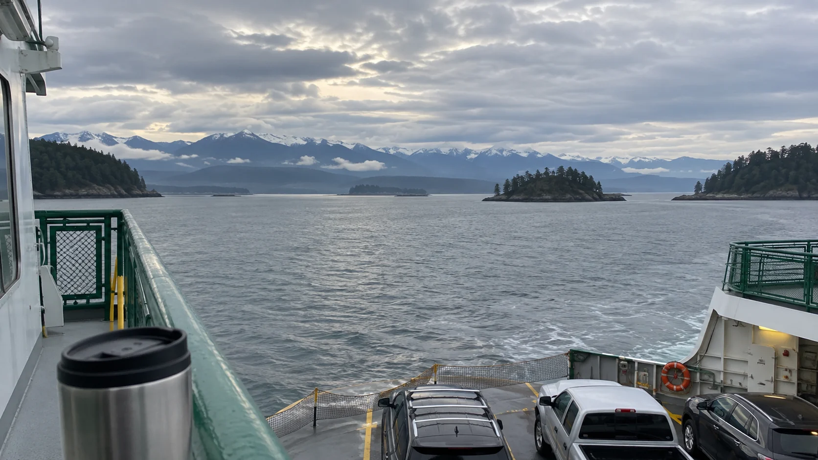 Road-trip vehicles and a travel mug aboard a ferry crossing Puget Sound toward the Olympic Mountains.