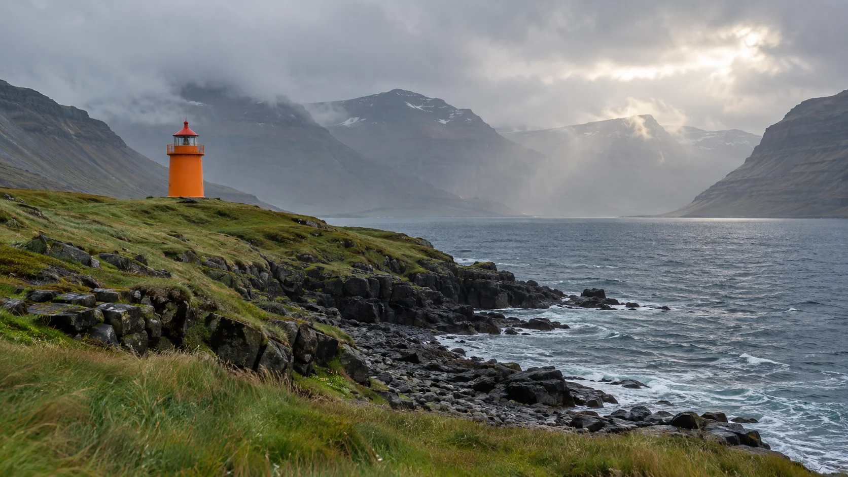 Orange Vattarnesviti lighthouse on a rocky Eastfjords cape beneath rain-wrapped mountains.