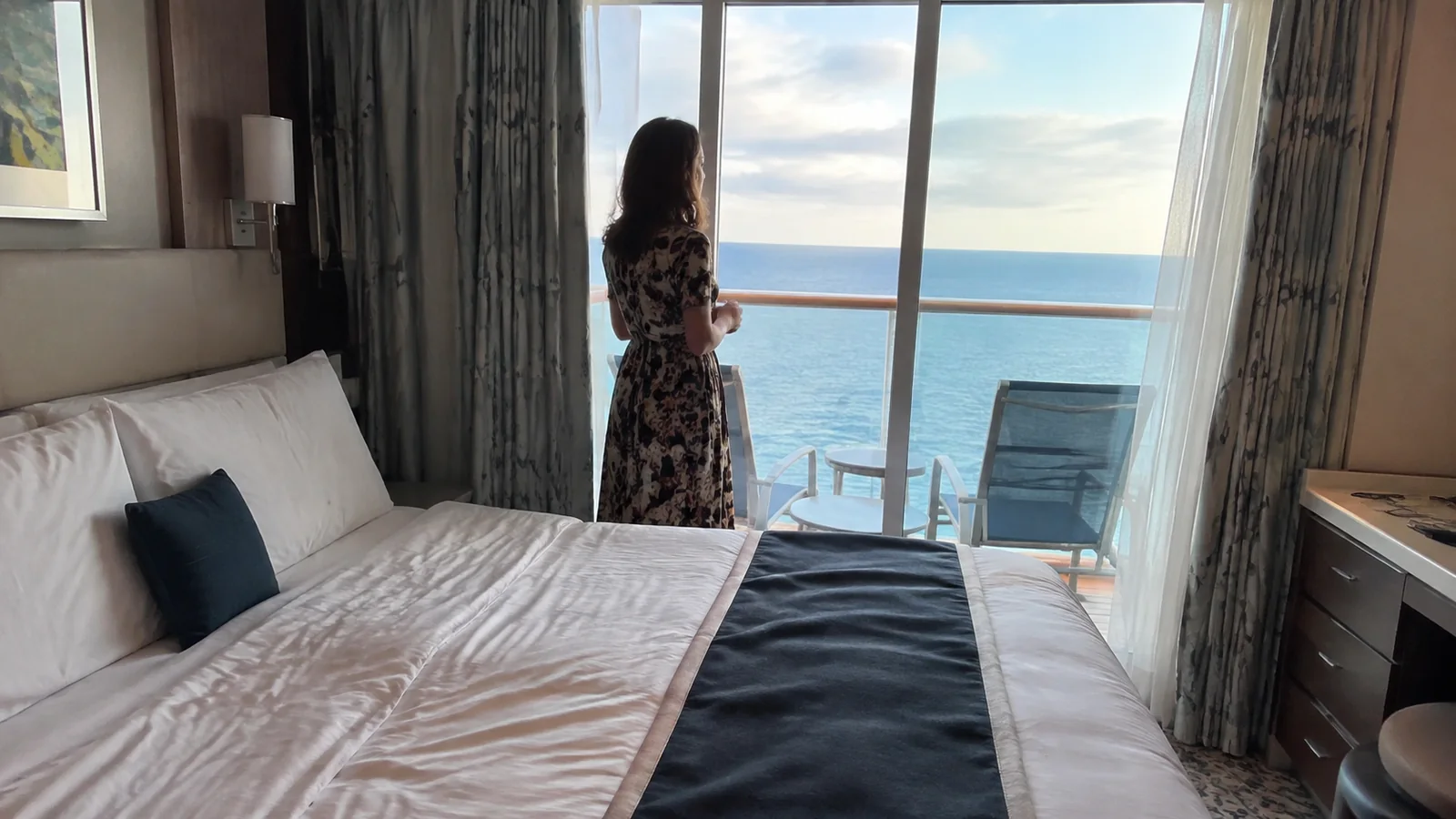 Woman standing beside balcony doors in an ocean-view ship cabin.