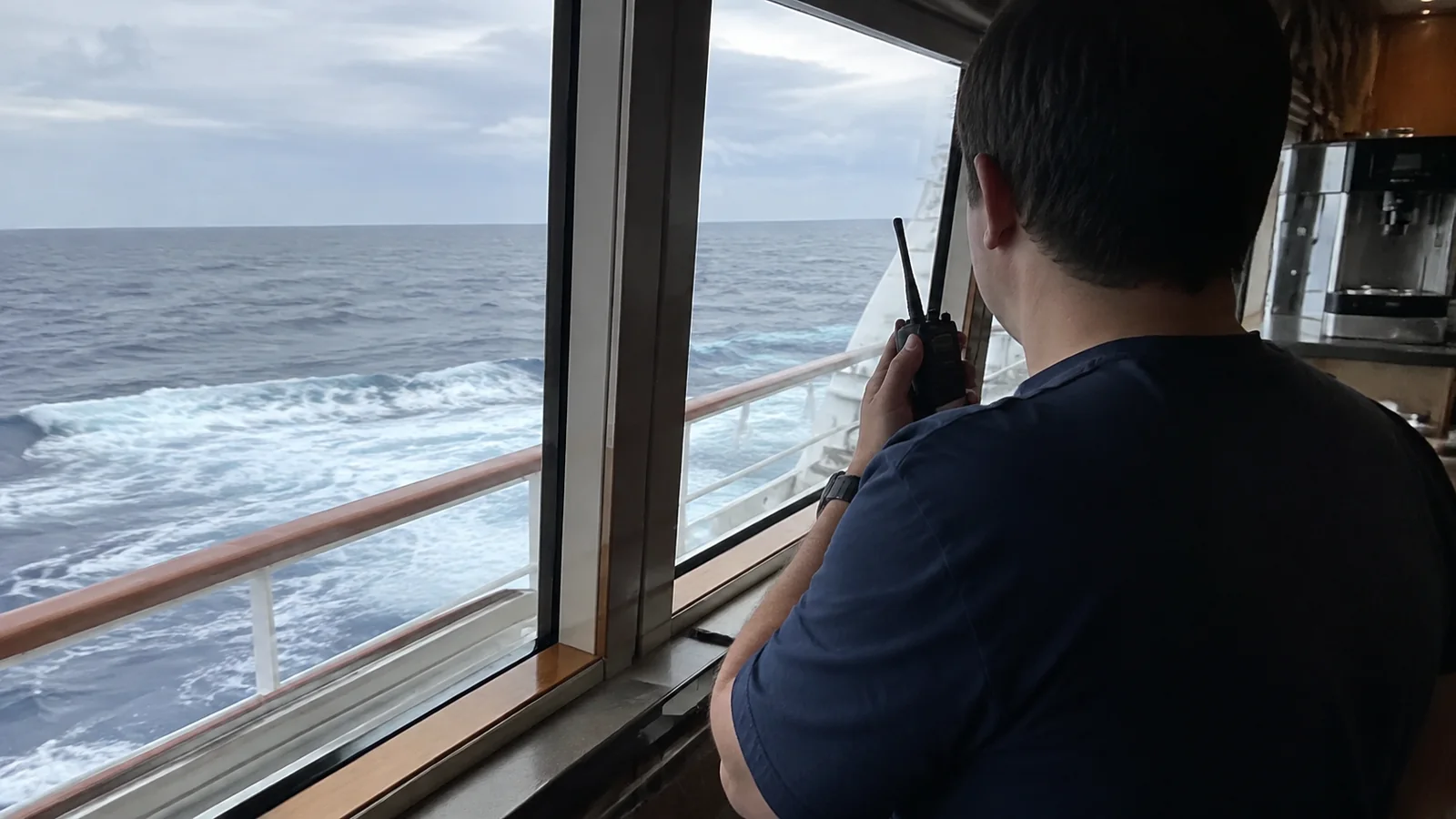 Man using a walkie-talkie beside a cruise ship window overlooking the wake