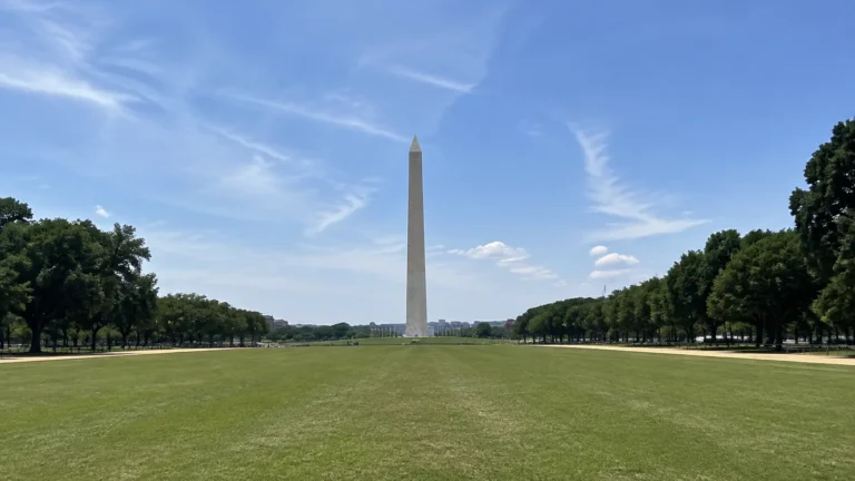 Tall pale obelisk rises above a broad lawn under blue skies.
