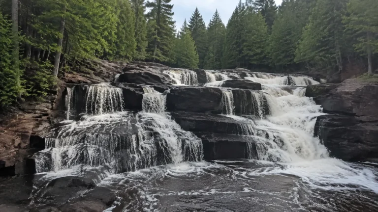 Broad stepped waterfall flows over dark rock ledges through evergreen forest.