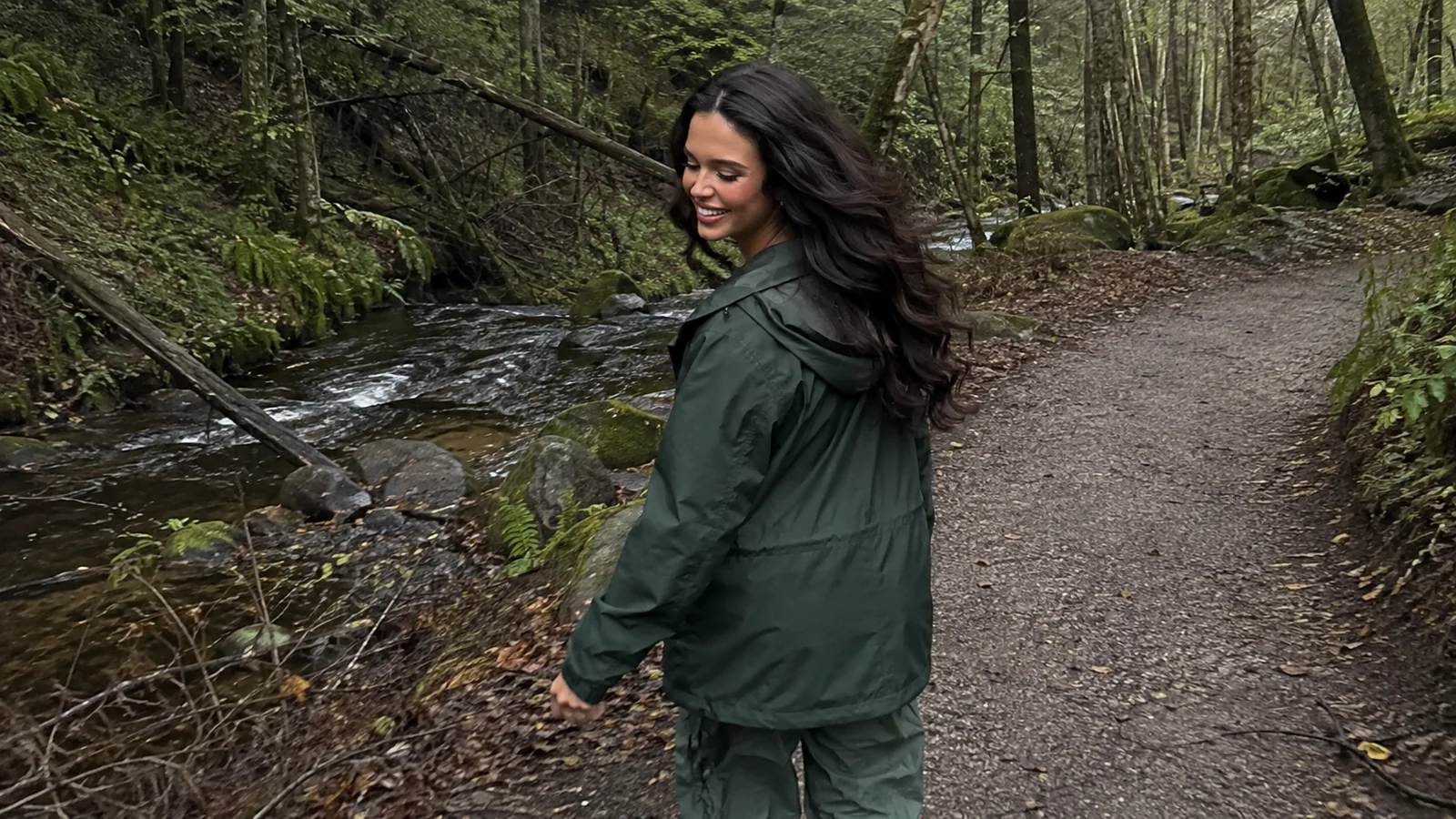 Woman in a green rain jacket walking a wooded riverside path.