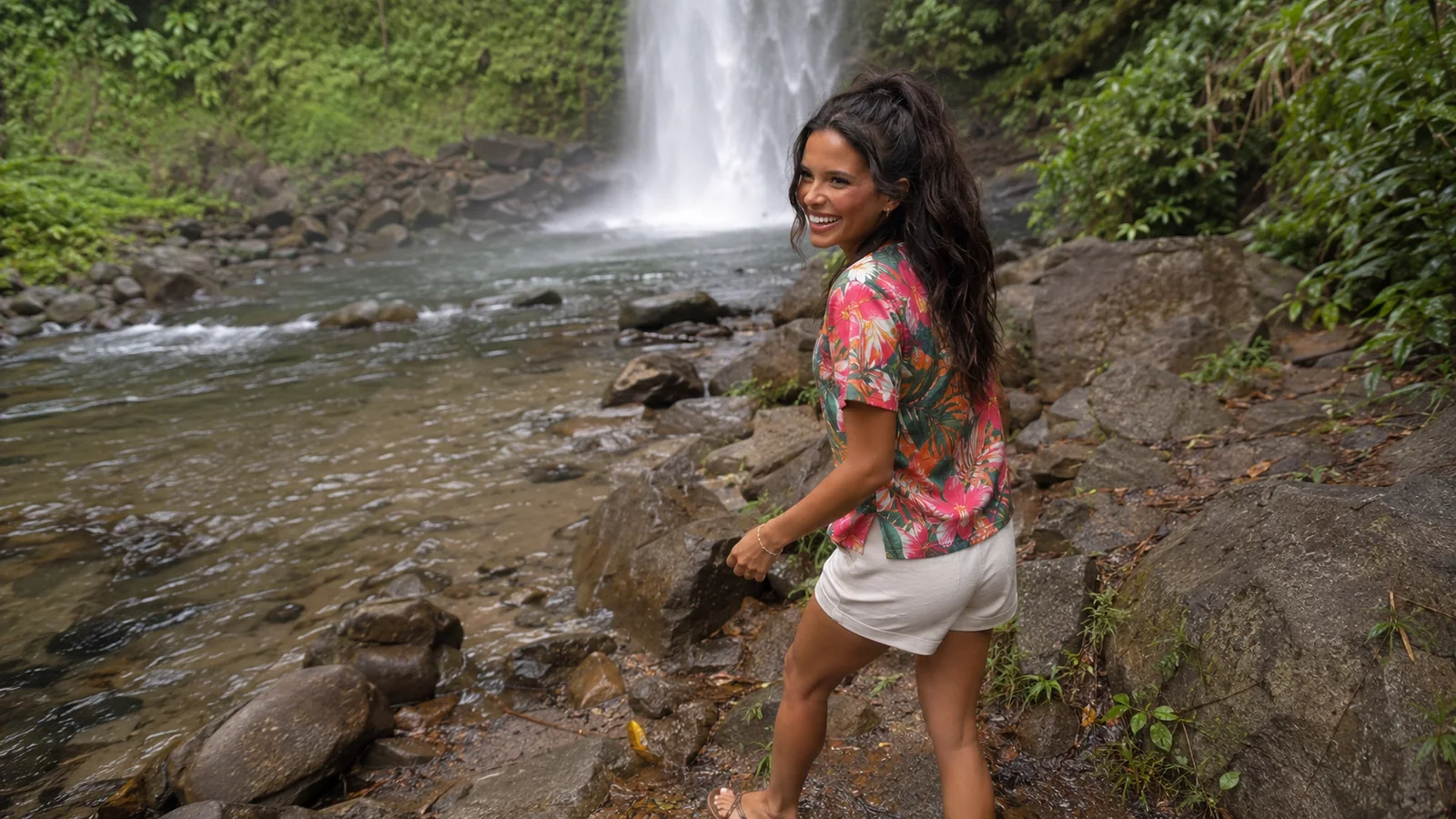 Woman walking along a rocky stream with a waterfall behind her