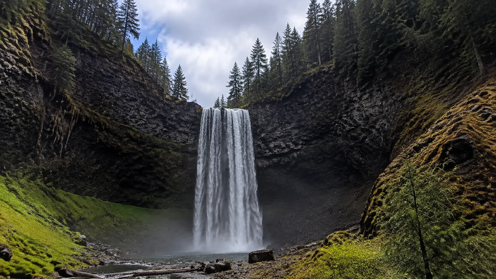 Tall narrow waterfall drops between steep forested rock walls.