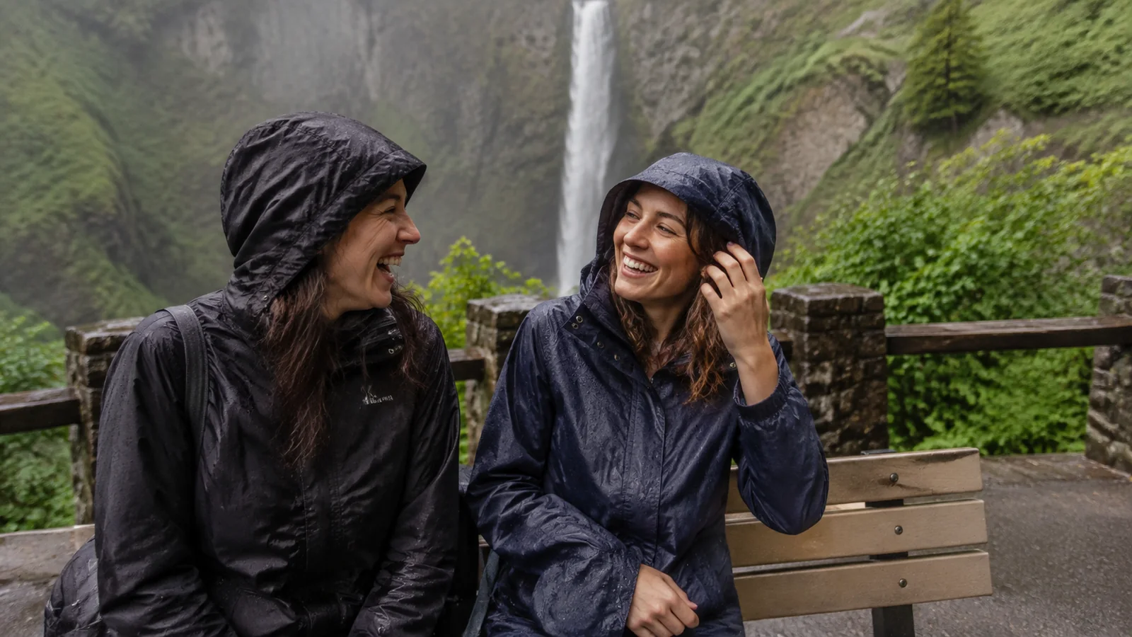 Two travelers in hooded rain jackets smiling beside a waterfall viewpoint.