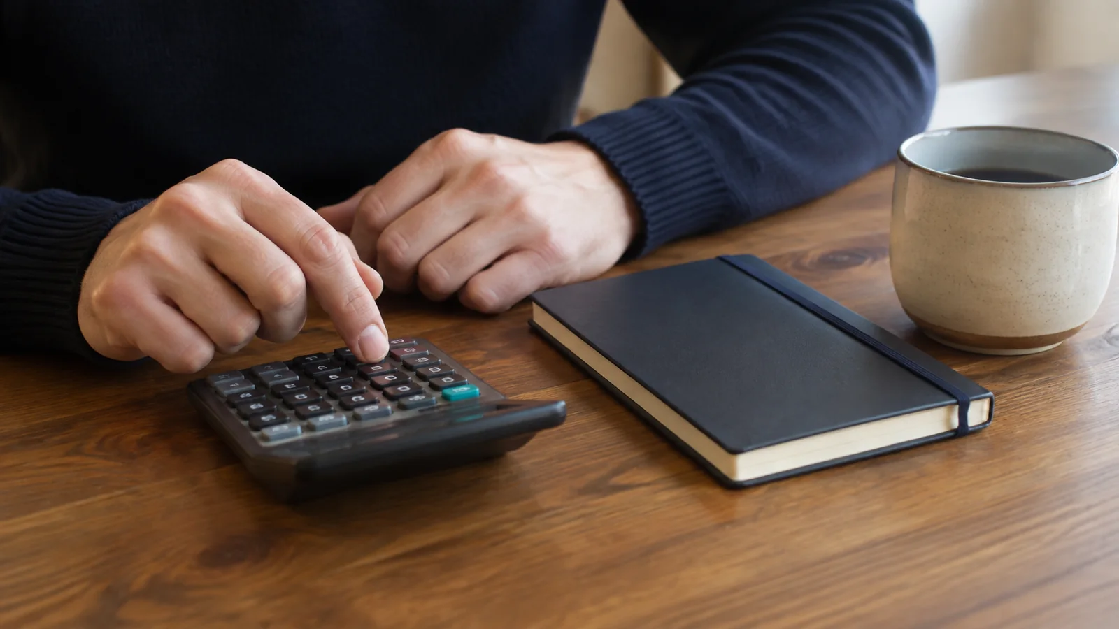 Hands operate a calculator beside a notebook and mug.