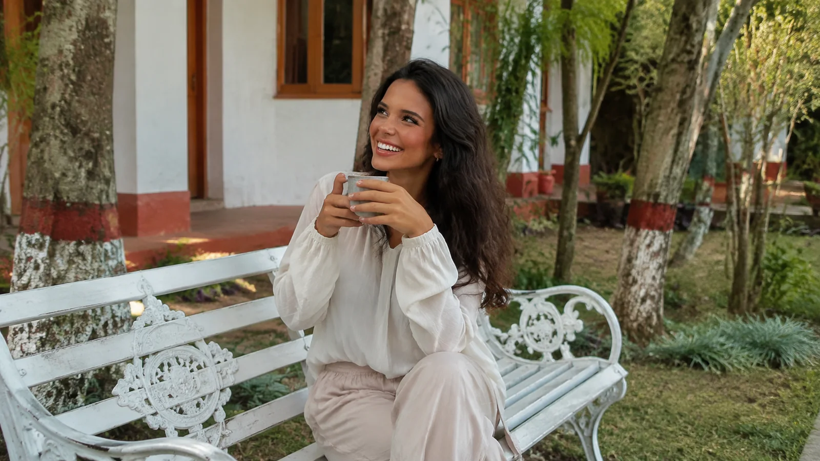 Woman holds a cup while seated on a white garden bench.