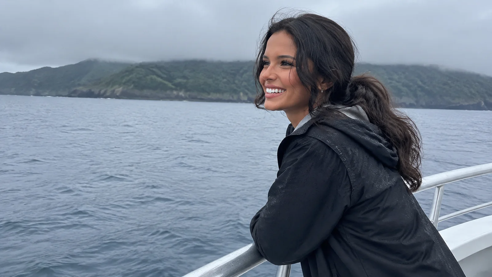 Smiling woman in a rain jacket at a boat railing overlooking misty mountains