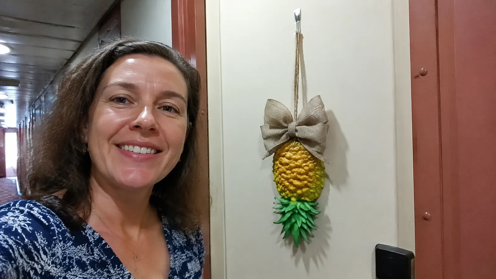 Smiling woman standing beside an upside-down pineapple decoration on a cruise cabin door