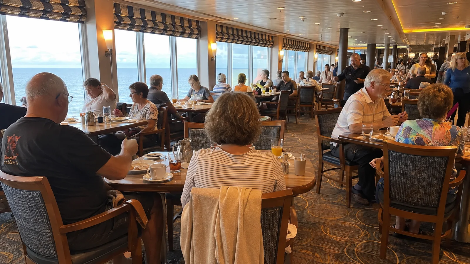 Crowded cruise ship dining room with passengers seated at tables beside large windows